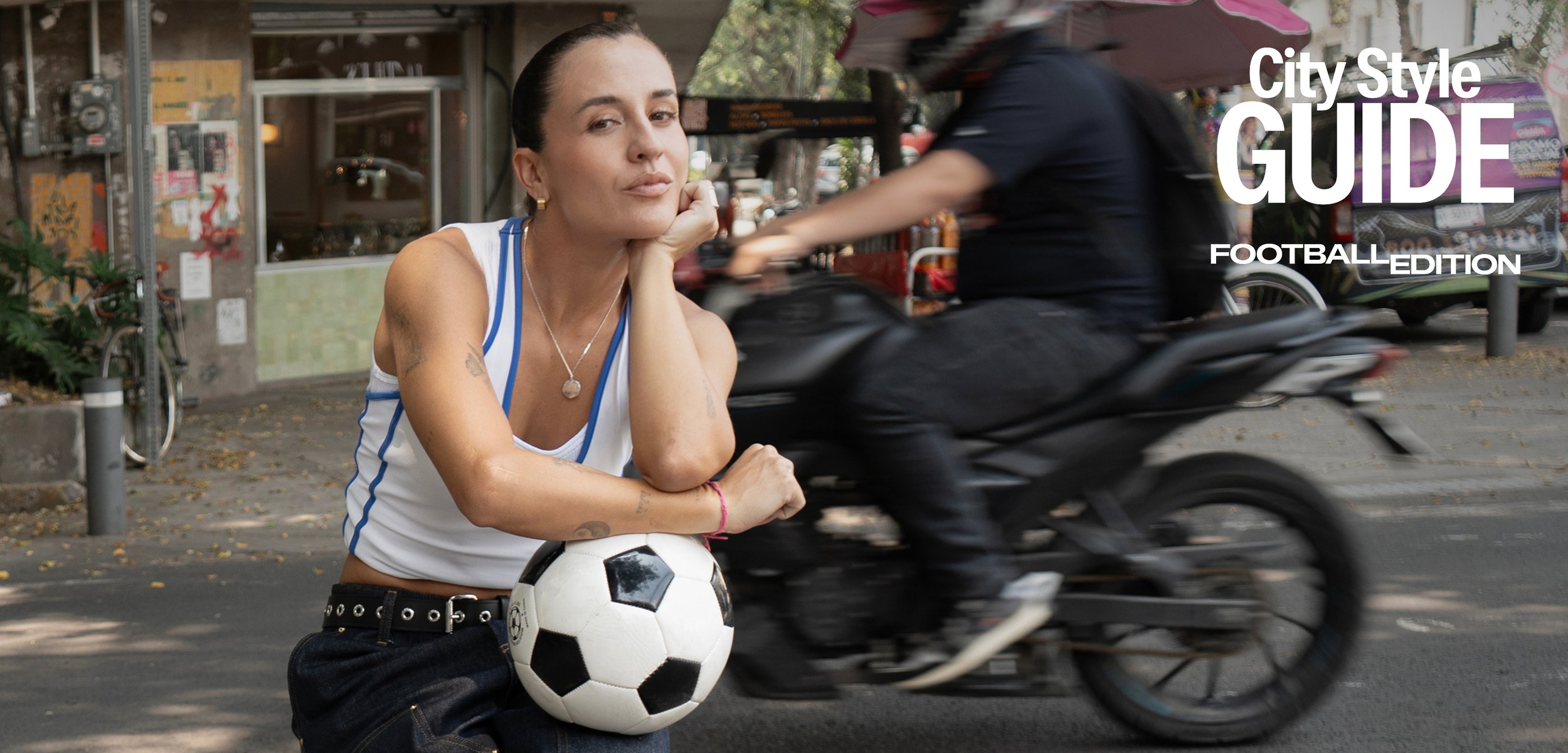 Femeie în haine sportive casual sprijină brațul pe o minge de fotbal în timp ce un motociclist trece pe o stradă din oraș.