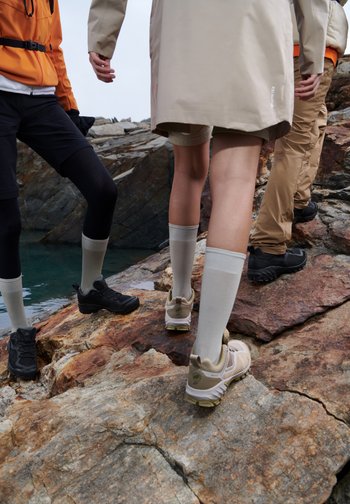 Drei Personen gehen auf felsigem Terrain neben einem Gewässer und tragen Outdoor-Ausrüstung, einschließlich Shorts, Socken und Wanderschuhen.