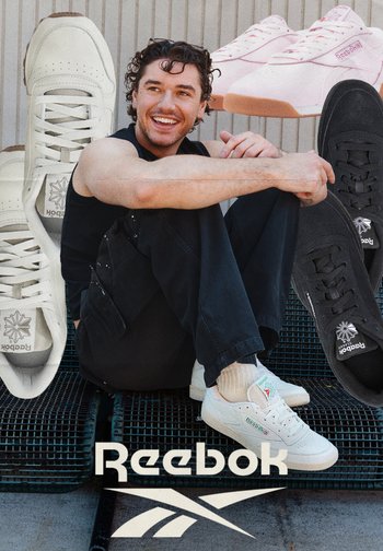 Junger Mann in schwarzer Kleidung, der lächelt, während er sitzt, umgeben von großen weißen, pinken und schwarzen Reebok-Sneakers und dem Reebok-Logo darunter.