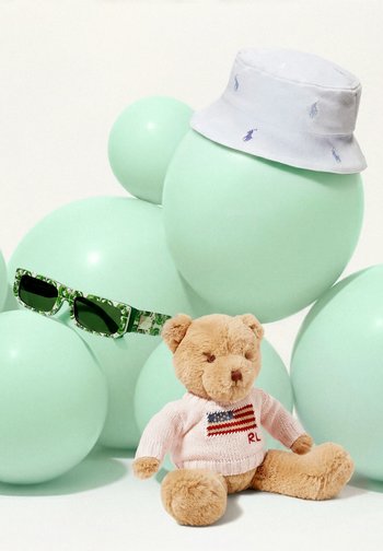 Plišasti medvedek, oblečen v roza pulover z ameriško zastavo, mint zelene balone, sončna očala in belo skodelico s kapo z vezenim logotipom.