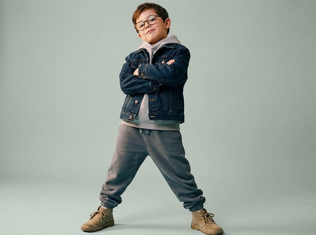 Jeune garçon portant des lunettes, une veste en jean, un sweat à capuche gris et un pantalon de survêtement, se tenant avec confiance, bras croisés et jambes écartées, sur un fond uni.