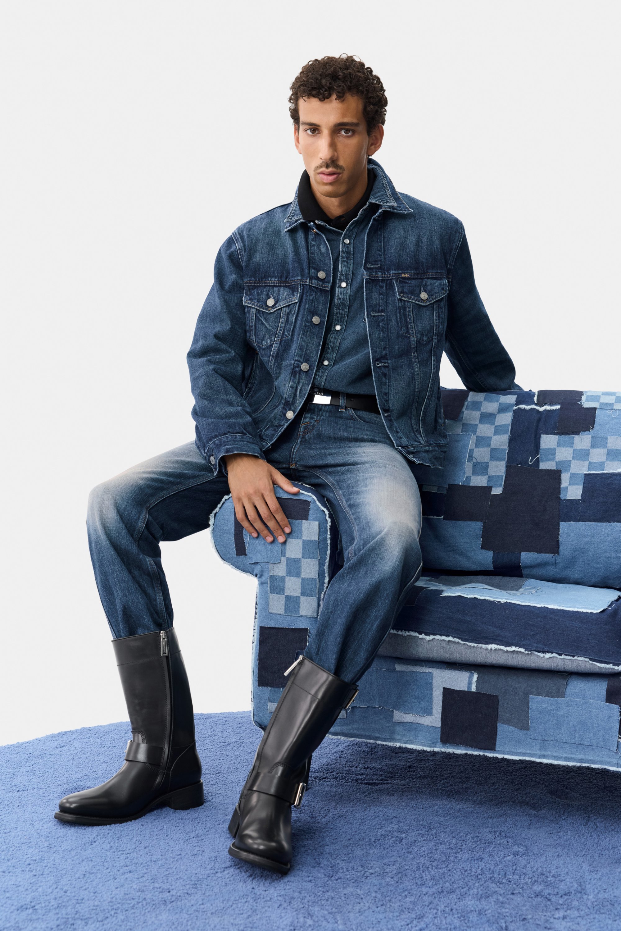Donkerblauwe spijkerjas en spijkerbroek, gecombineerd met zwarte leren laarzen met ritsen en zilveren accenten, zittend op een patchworkbank.
