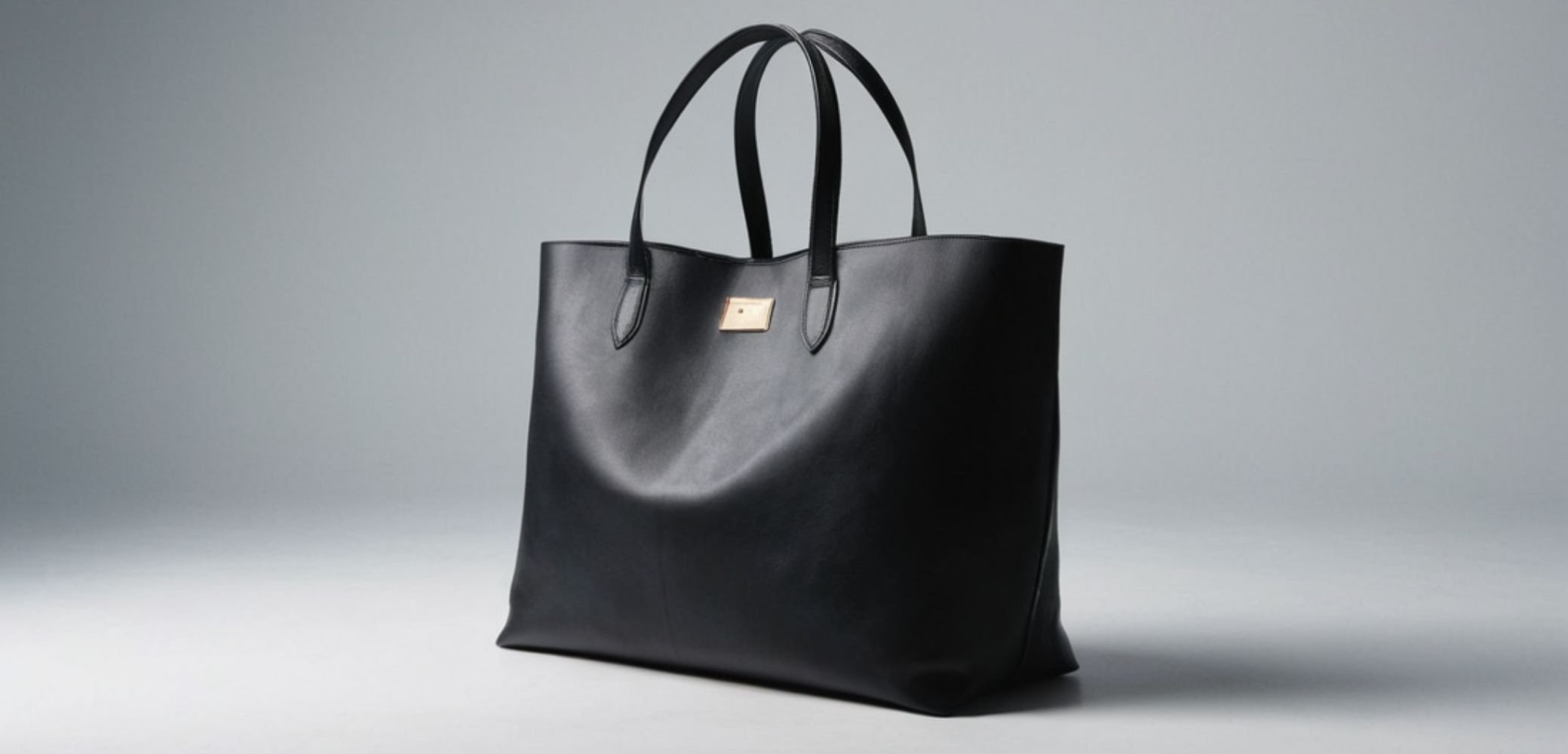 Duża czarna skórzana torba na ramię z dwoma uchwytami i małym złotym emblematem z przodu, na neutralnym szarym tle.