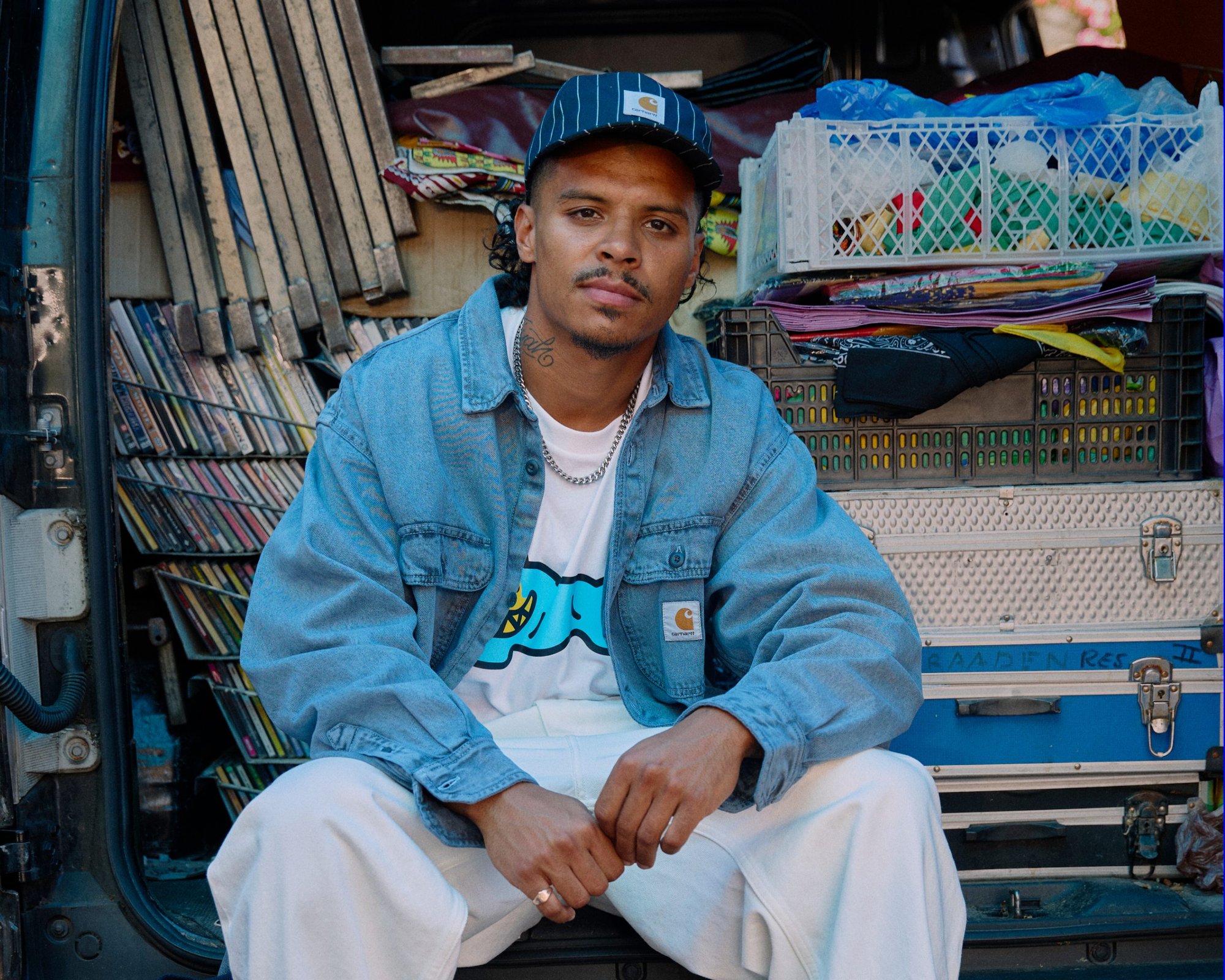 Un jeune homme portant une veste en denim bleu et un pantalon blanc est assis devant une fourgonnette remplie de boîtes colorées et de cassettes, portant une casquette noire.
