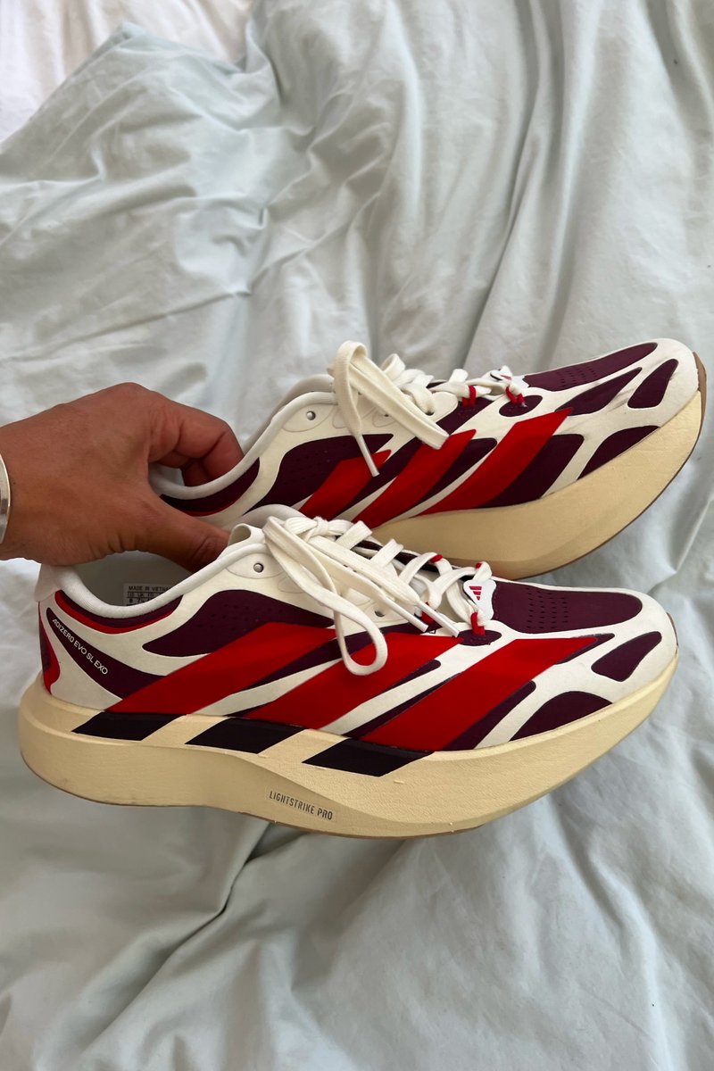 Mano sosteniendo un par de zapatillas para correr blancas, rojas y granate con suelas gruesas beige sobre una superficie de tela azul claro.