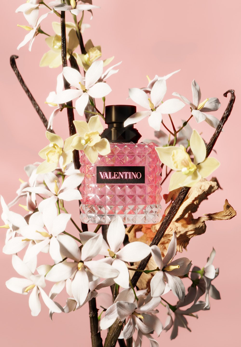 Flacon de parfum Valentino rose avec bouchon noir entouré de fleurs blanches et jaune pâle sur un fond rose clair.