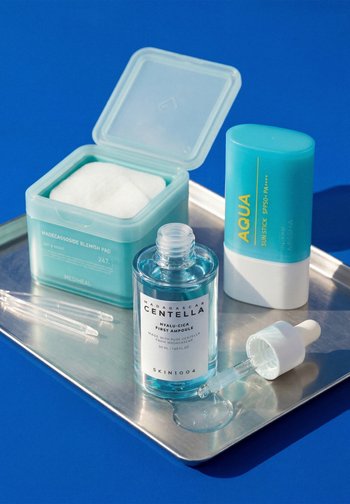 Ampolla per la cura della pelle azzurra trasparente con contagocce, tamponi per imperfezioni in contenitore turchese aperto e stick solare aqua su vassoio d'argento su sfondo blu.