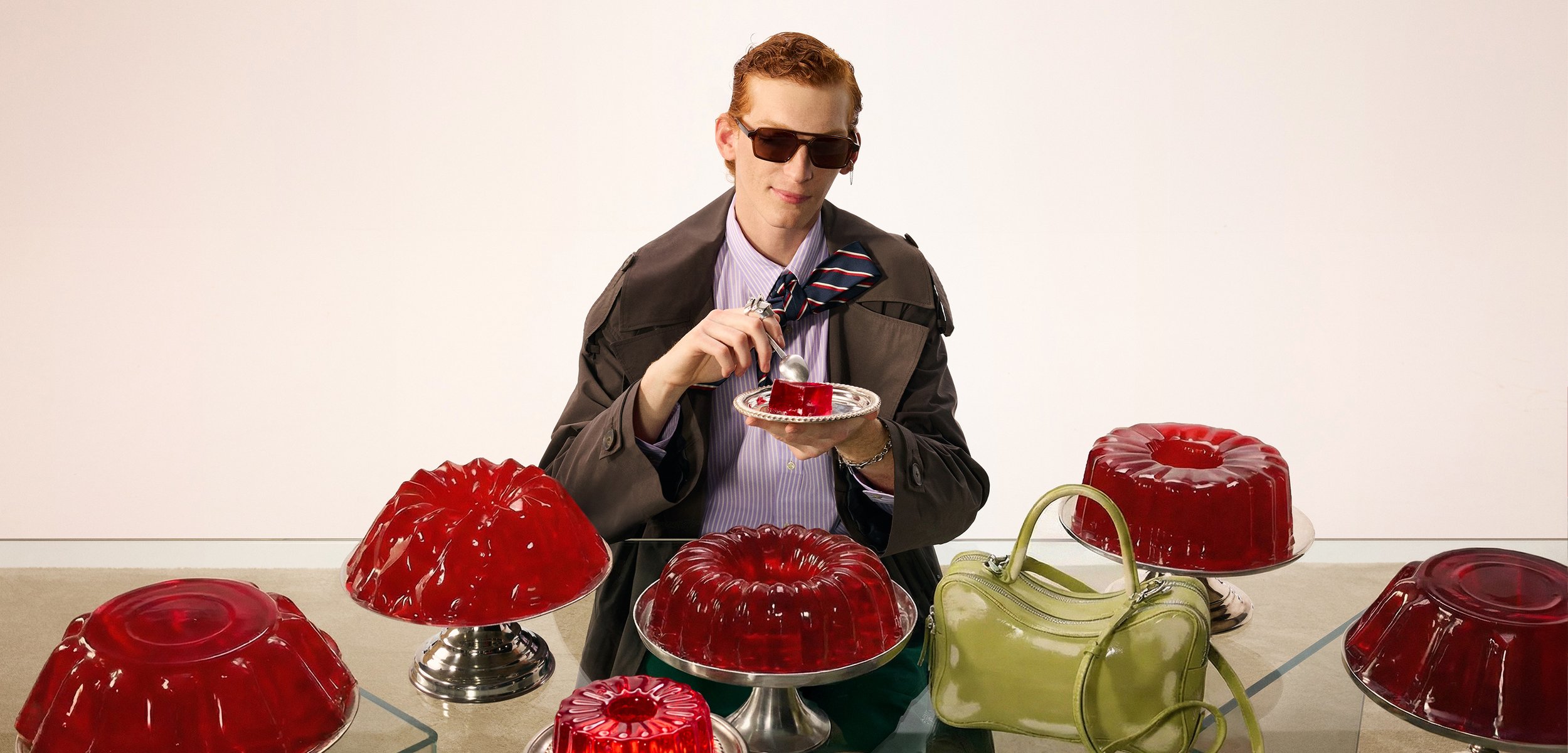 Fiatal férfi napszemüvegben és kabátban egy villát és egy tányért tart piros zselével, öt nagy piros zselékupac és egy zöld kézitáska veszi körül.