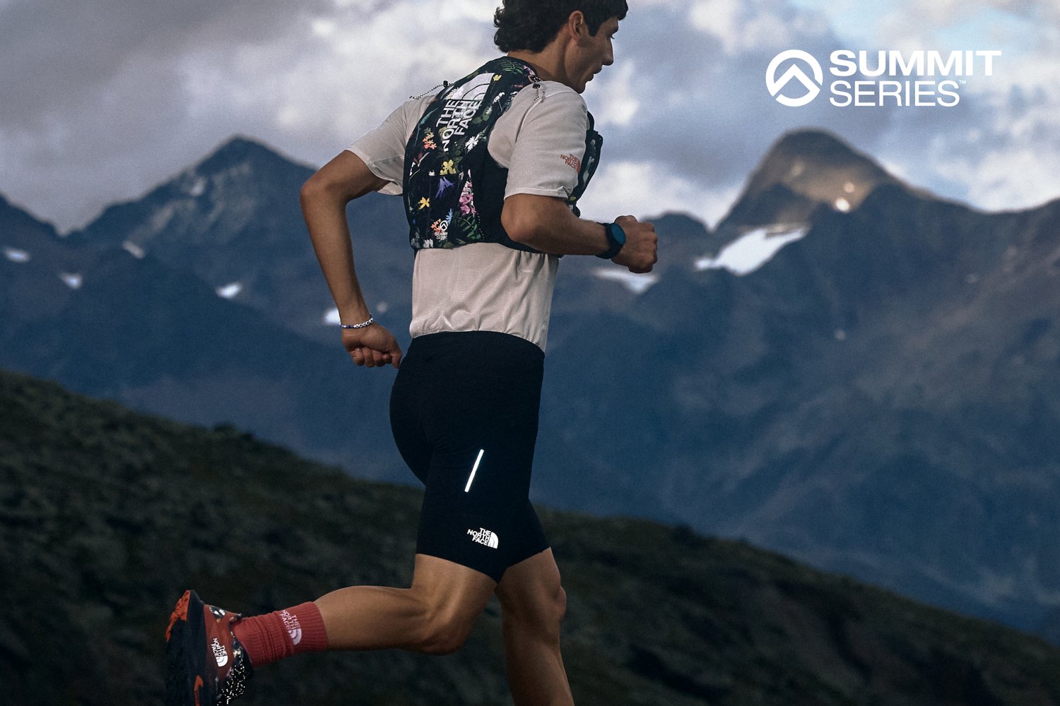 Corredor de trail con un chaleco de hidratación de patrón floral, camiseta gris, pantalones cortos negros con una franja reflectante y calcetines rojos, en un terreno montañoso rocoso.