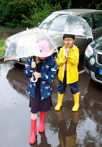 Deux enfants portant des imperméables et des bottes tiennent des parapluies transparents tout en se tenant dans un parking mouillé avec des flaques d'eau et des voitures garées.