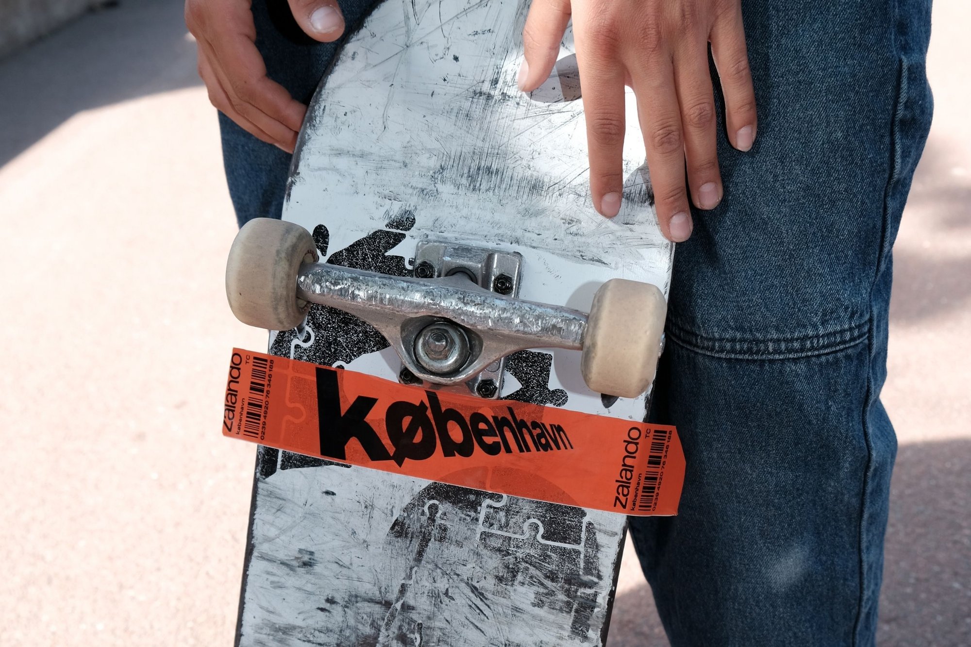 Skateboard med ett vitt, märkt bräda; silverfärgade truckar; beige hjul; har en orange klisterlapp märkt "københavn" och "zalando."