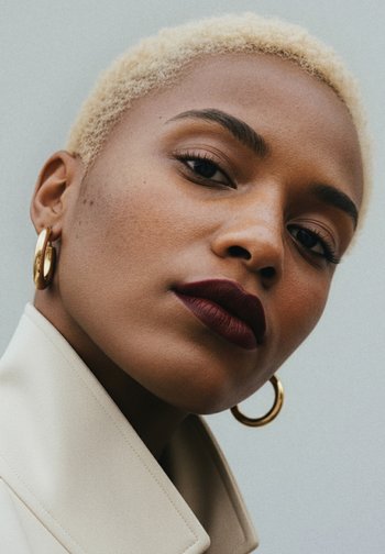 Retrato en primer plano de una persona con cabello corto y rubio, y labios pintados de un tono oscuro, que lleva una chaqueta color crema y grandes pendientes de aro dorados.