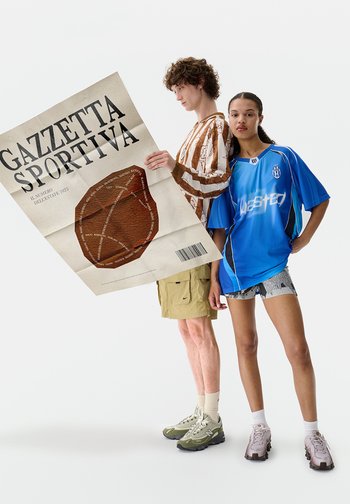 Dois jovens adultos estão de costas um para o outro; um lê um grande jornal "Gazzetta Sportiva", enquanto o outro veste uma camisola desportiva azul e calções.