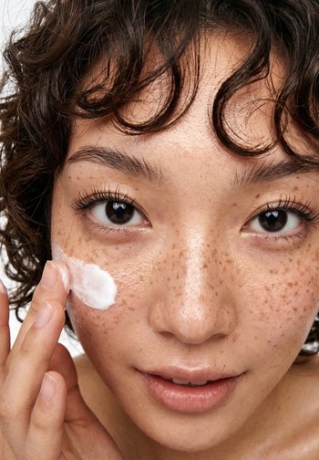 Personne aux cheveux bouclés et aux taches de rousseur appliquant une crème blanche sur la joue avec les doigts, regardant droit devant.