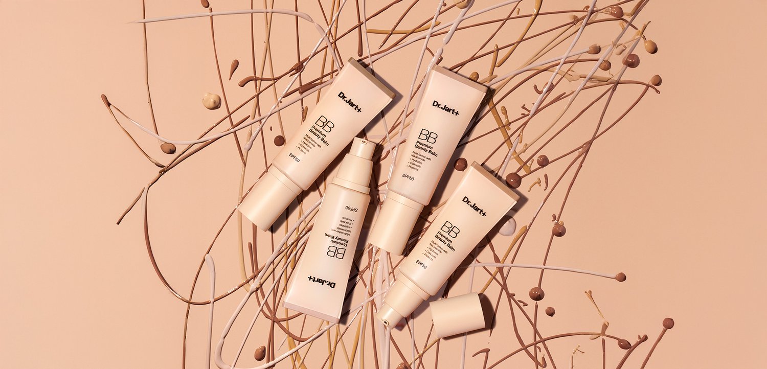 Fünf beige und hellbraune Tuben der Dr. Jart+ BB-Creme mit geprägten Etiketten, angeordnet zwischen dünnen, geschwungenen beigen und braunen Linien vor einem rosa Hintergrund.