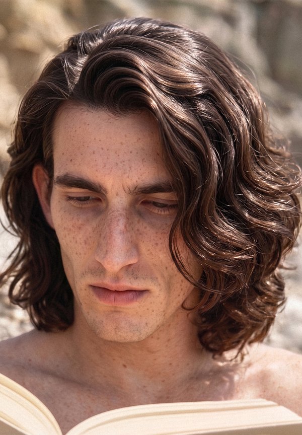Jeune homme aux longs cheveux bruns ondulés et taches de rousseur, regardant vers le bas et lisant un livre en plein air.