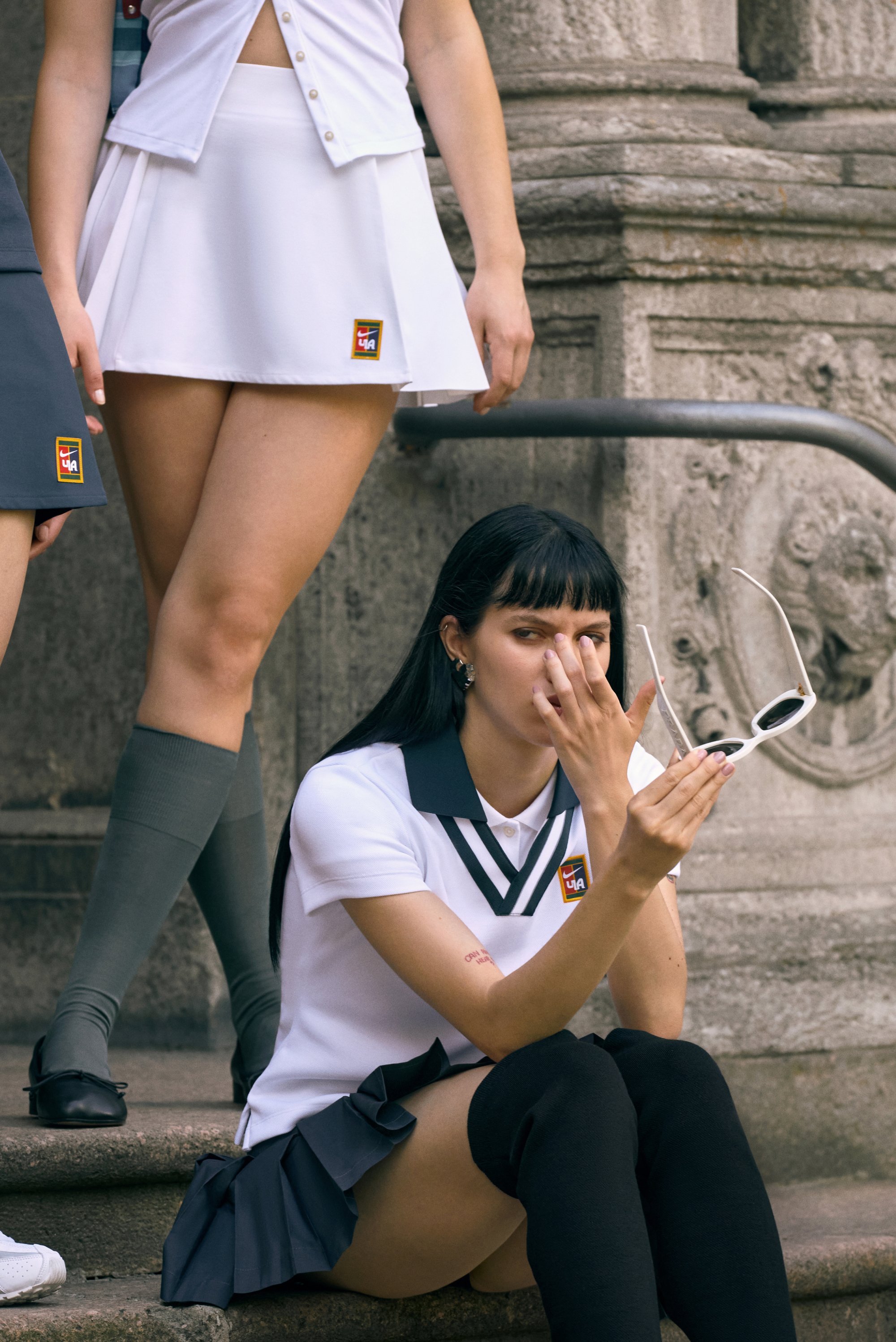 Due giovani donne in gonna e calze al ginocchio posano su gradini di pietra. Una è seduta, mentre aggiusta degli occhiali da sole bianchi, mentre l'altra sta in piedi nei pressi.