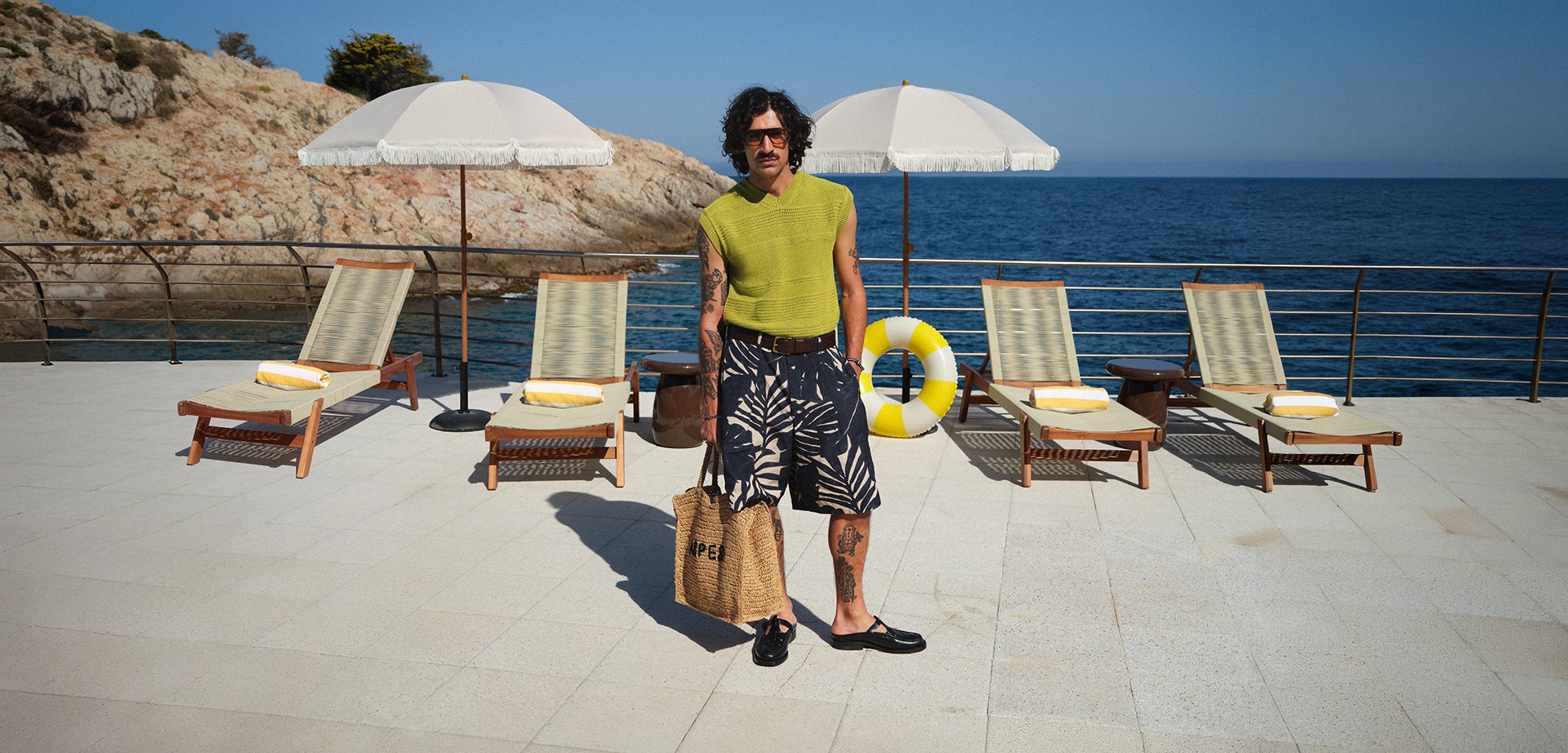 Homme en haut sans manches vert et short à motifs se tient sur une terrasse ensoleillée avec des chaises longues en bois, des parasols et l'océan en arrière-plan.