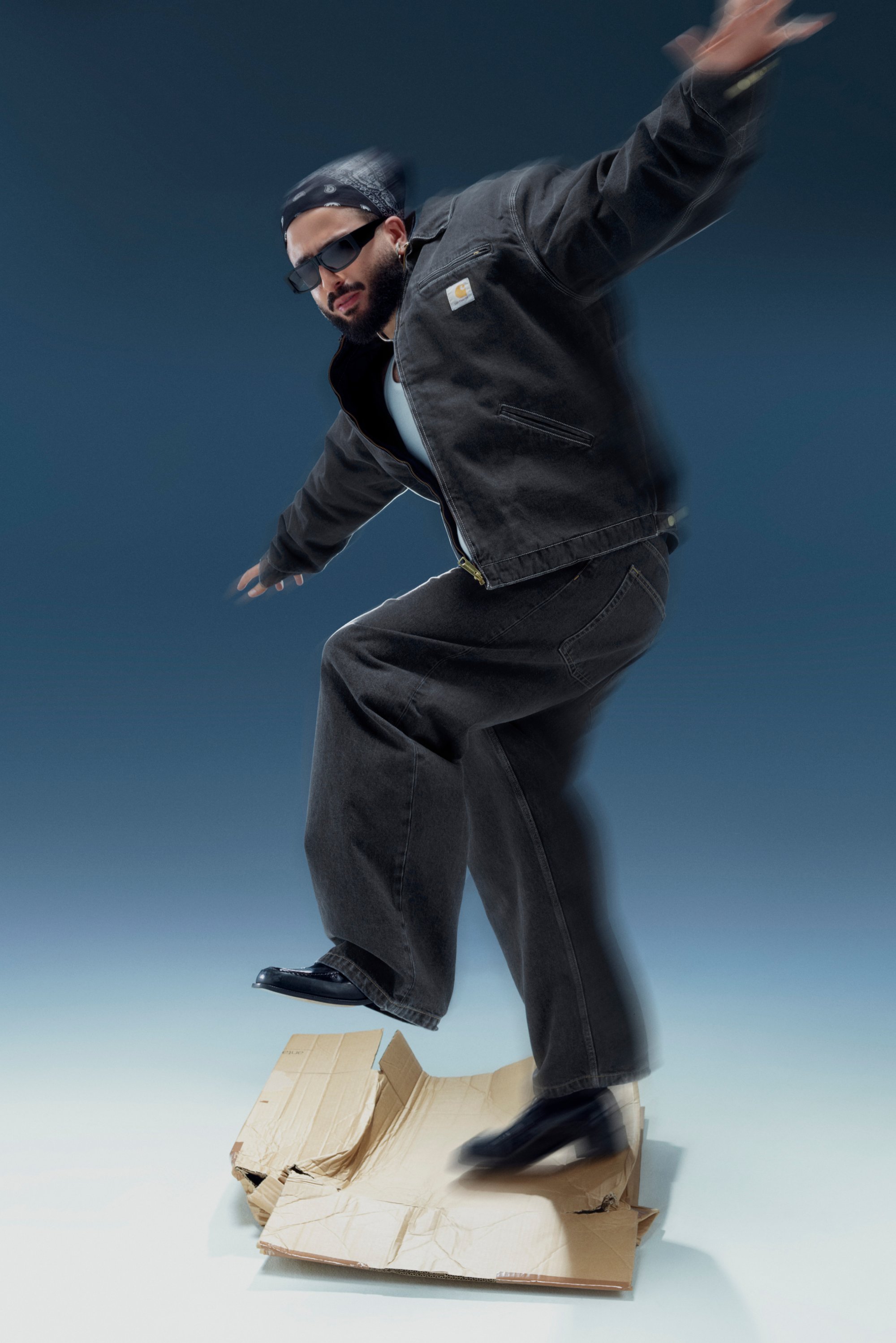 Een man in een zwarte denim outfit en zonnebril balanceert op een afgevlakte kartonnen doos, wat dynamisch lijkt tegen een vervaagde achtergrond.