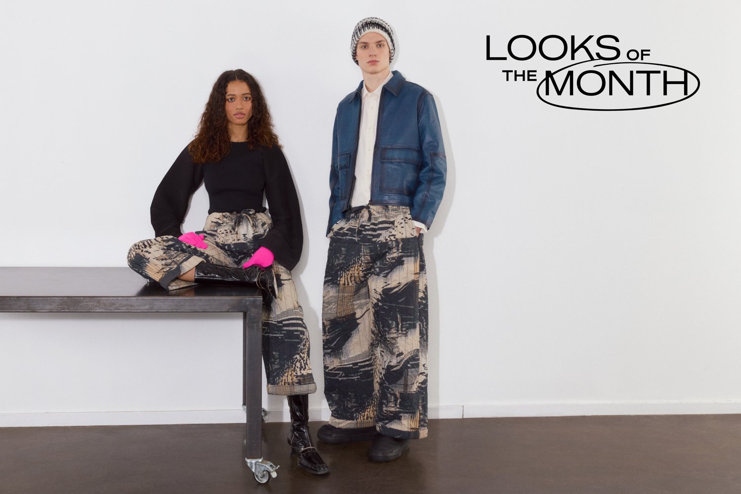 Una mujer con cabello rizado está sentada en una mesa, vistiendo pantalones negros y estampados con guantes rosas. Un hombre está de pie a su lado con una chaqueta azul y pantalones estampados.
