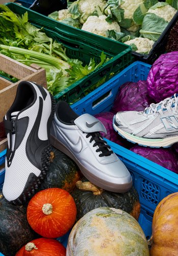 Fekete és ezüst sportcipők texturált felülettel és fűzős dísszel, különböző tökökön elhelyezve, zöld és lila zöldségek között ládákban.