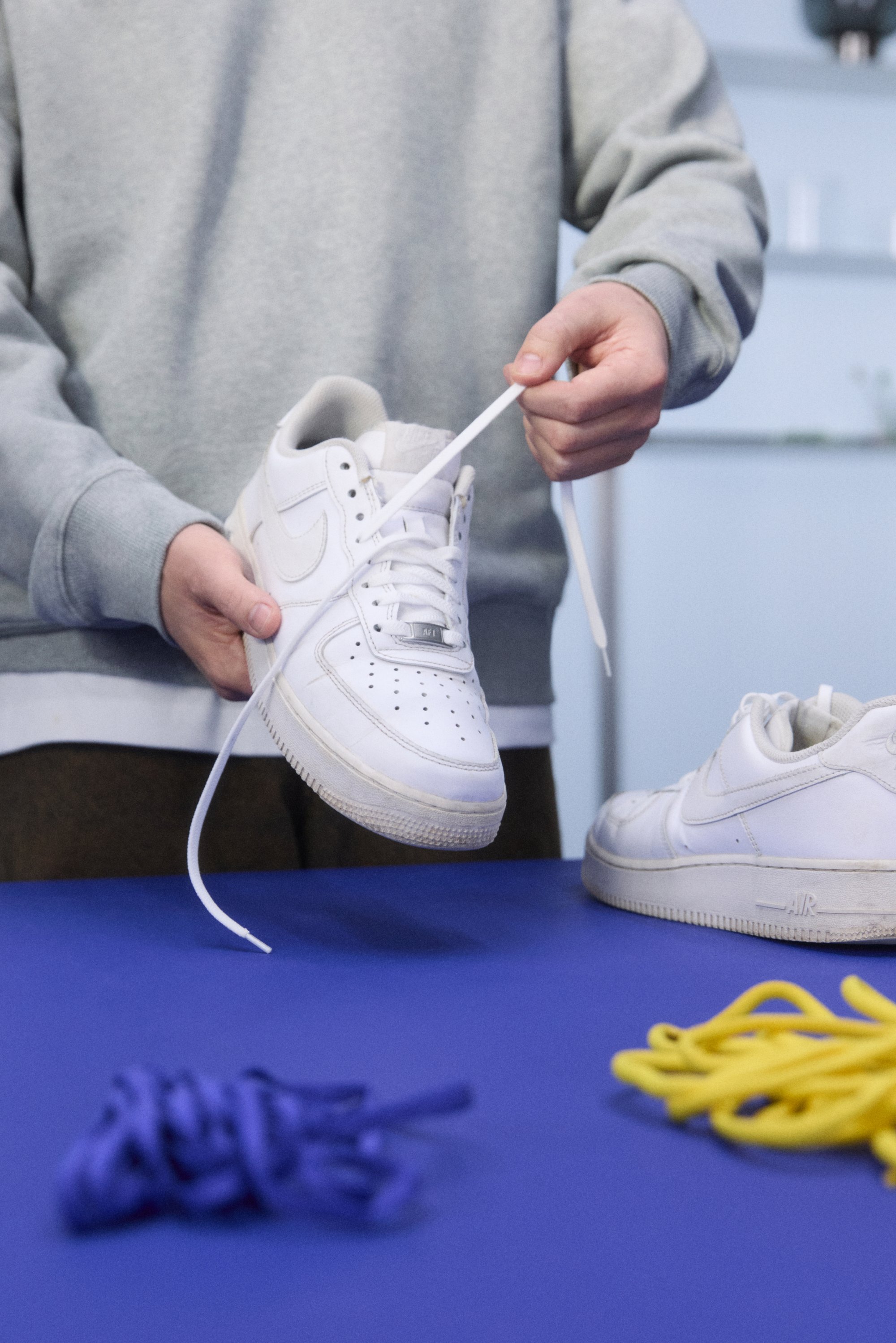 Valkoinen Nike Air Force 1 -lenkkari pidetään kädessä, esittäen nahkamateriaalia, rei'itetty kärki ja litteät nauhat. Keltaiset ja purppurat nauhat sinisellä pinnalla.