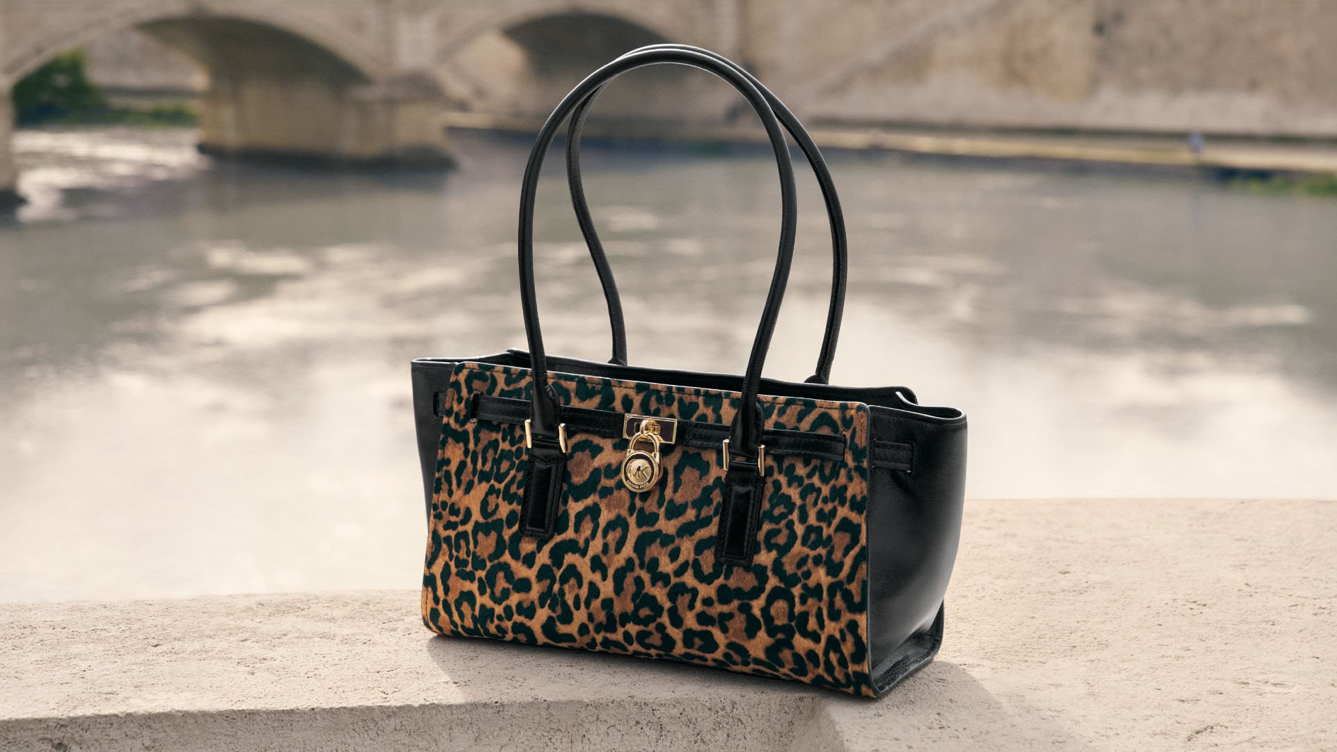 Geantă de mână cu model leopard maro și negru, cu accente din piele neagră, două mânere, accesorii aurii și formă rectangulară.