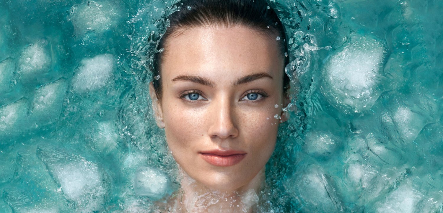 Een close-up van het gezicht van een vrouw, ondergedompeld in turkooizen water, omringd door luchtbelletjes en ijsblokjes, met de nadruk op haar heldere huid en blauwe ogen.