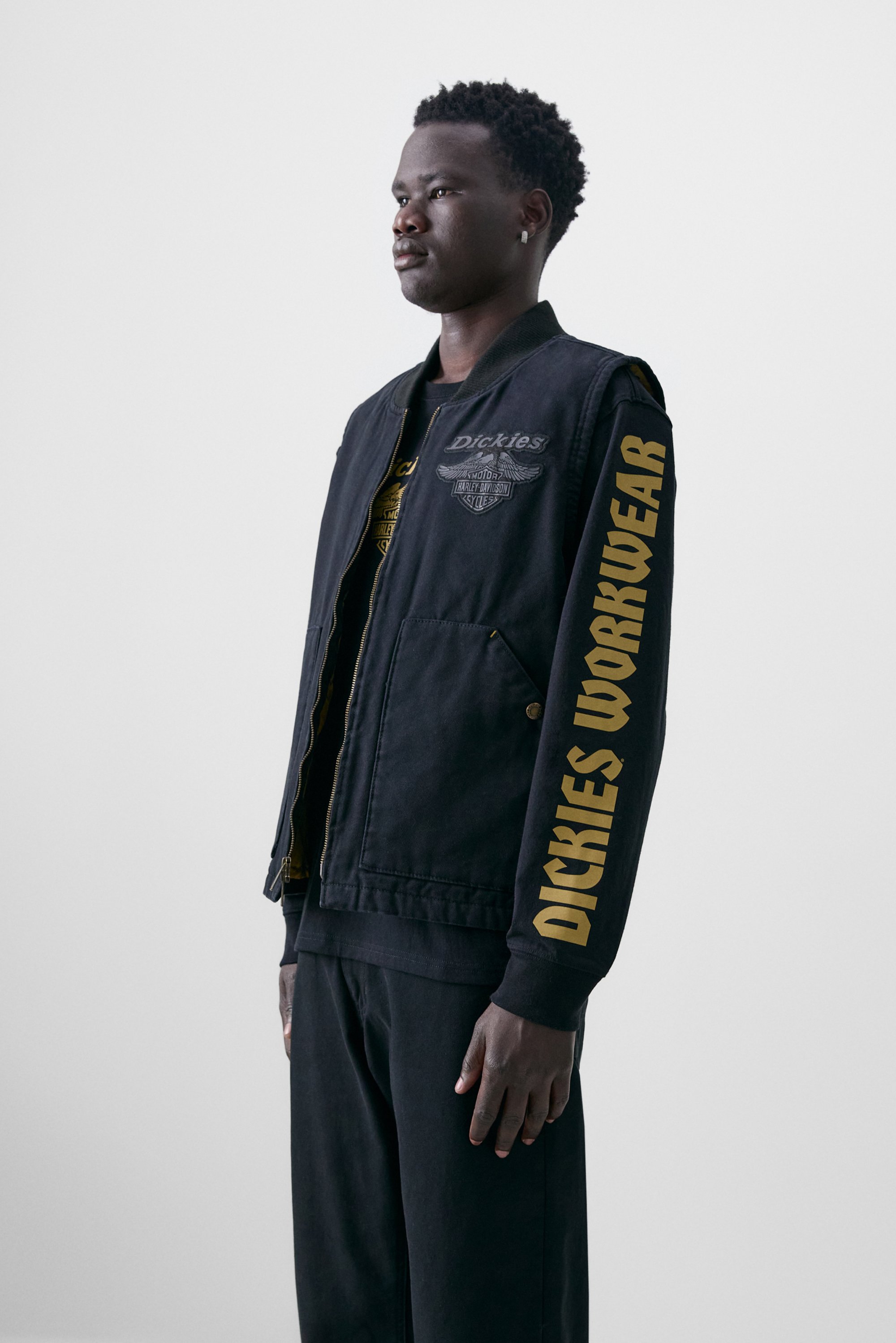 Persoon in een zwart jack met "Dickies" branding, gekleed in een zwart outfit, tegen een effen witte achtergrond.