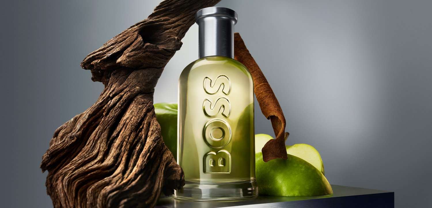 Bottiglia di vetro trasparente con tappo argento etichettata BOSS, circondata da fette di mela verde, legno secco e un bastoncino di cannella su una superficie grigia.