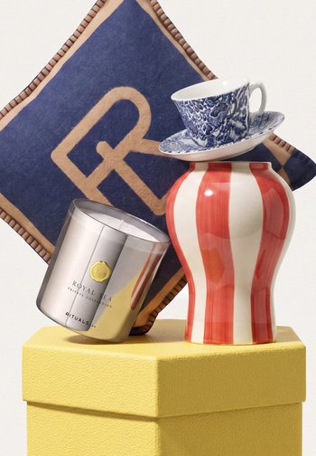 Coussin bleu avec motif doré derrière une bougie Royal Tea argentée, un vase rayé rouge et blanc, et une tasse à thé bleue décorée avec sa soucoupe sur une surface texturée jaune.