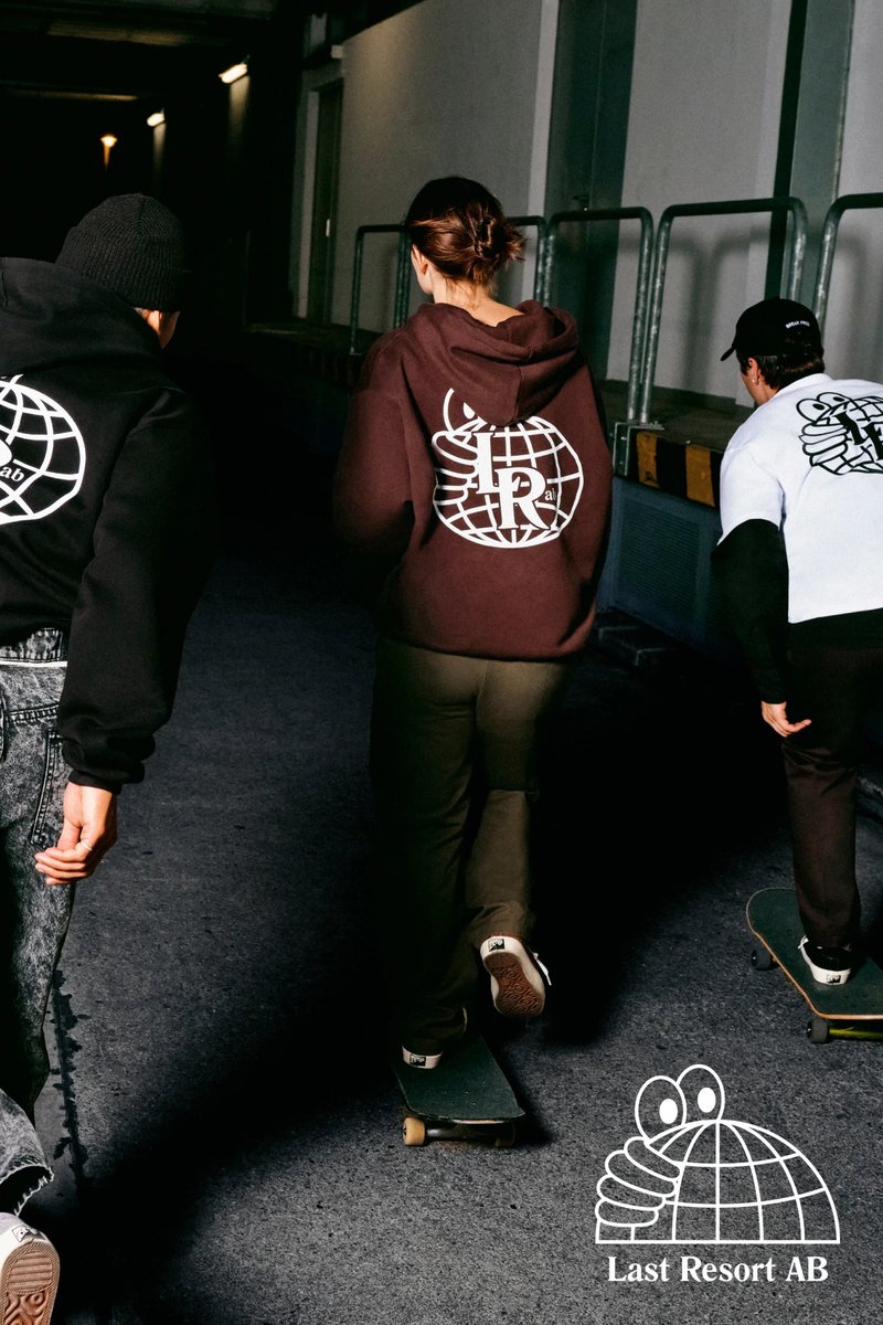 Tre individui in abbigliamento casual pattinano in un vicolo poco illuminato, mostrando felpe con cappuccio e una maglietta con un logo grafico.