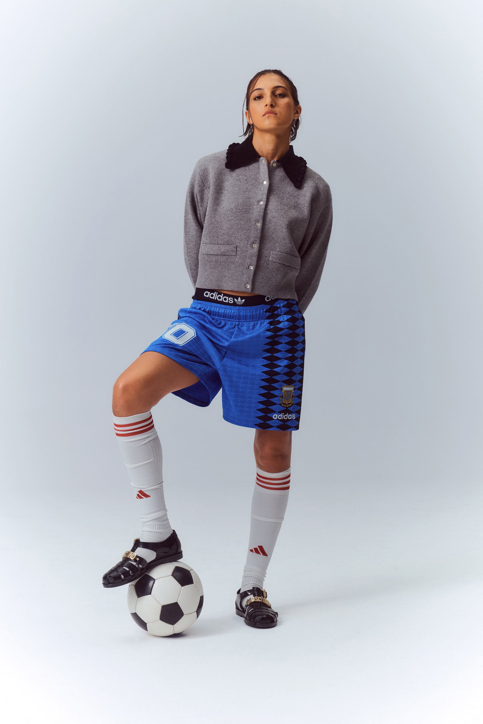 Una giovane donna con un cardigan grigio e pantaloni da calcio blu si erge con sicurezza con un piede su un pallone da calcio bianco e nero.