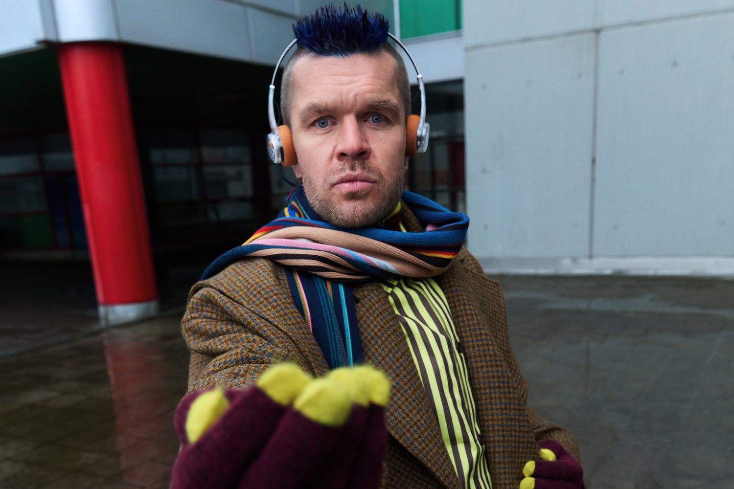 Mies, joka käyttää ruskeaa ruudullista takkia, raidallista huivia ja värikkäitä käsineitä, kuulokkeet päässään ja siniset hiukset, ojentaa käsineellistä kättään.