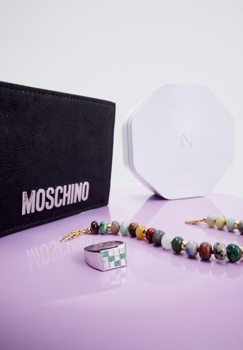 Schwarze Moschino-Geldbörse, eine weiße Box, ein farbenfrohes Perlenarmband und ein silberner Ring mit grünen Akzenten, die auf einer lavendelfarbenen Oberfläche angeordnet sind.