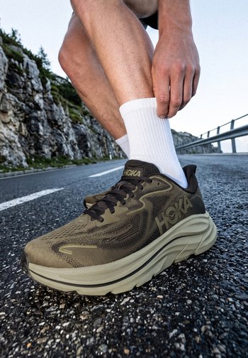 Person wearing white socks adjusting an olive green HOKA running shoe on a mountain road with rocky cliffs in the background.
