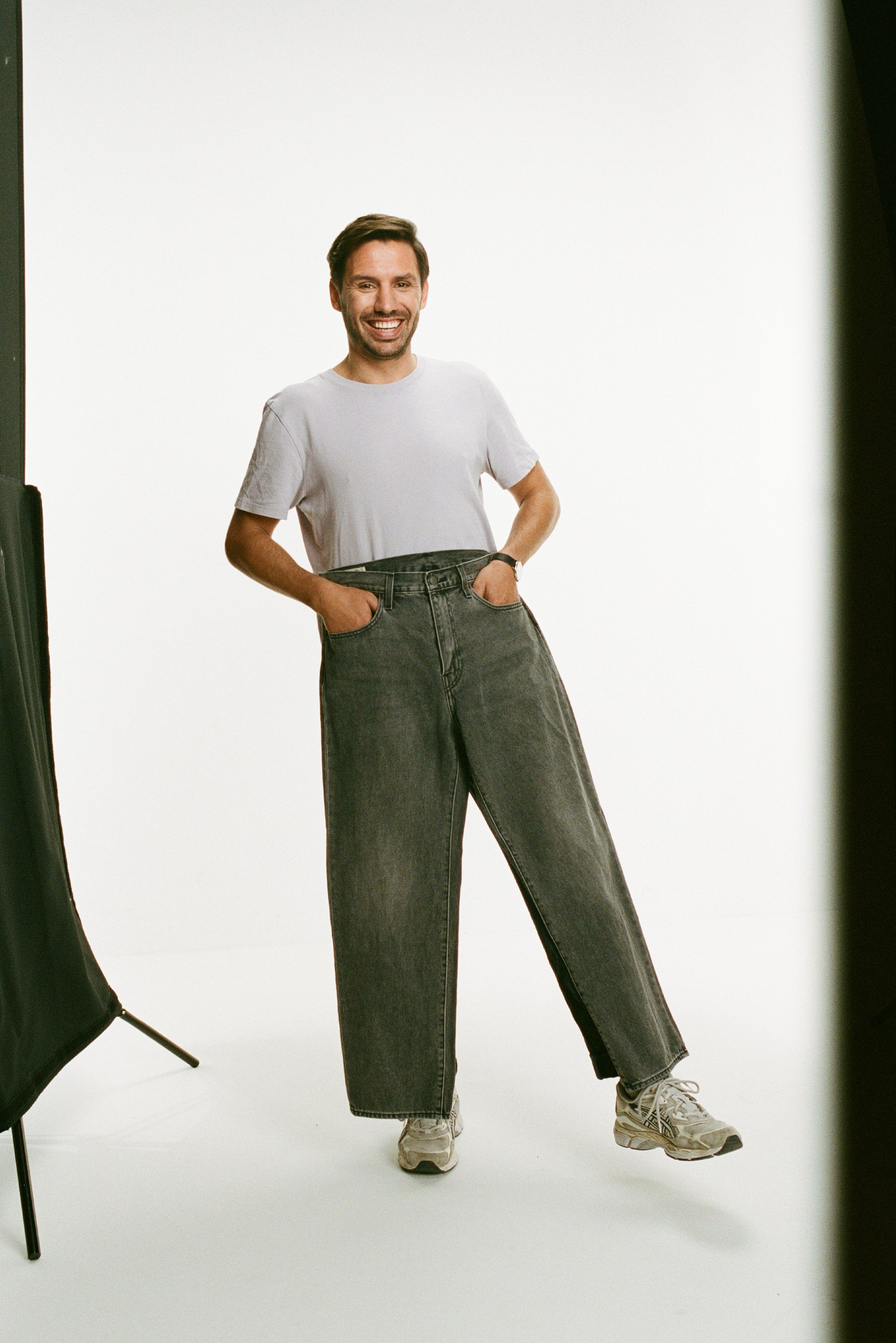 Uomo sorridente in una maglietta chiara sta in piedi con le mani sui fianchi, indossando jeans grigi oversize e scarpe da ginnastica, su uno sfondo neutro.