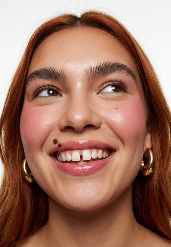 Vrouw met rood haar, gouden oorringen, glanzende lippen, een schoonheidsvlek boven haar lip en groene eyeliner die lacht en naar links kijkt.