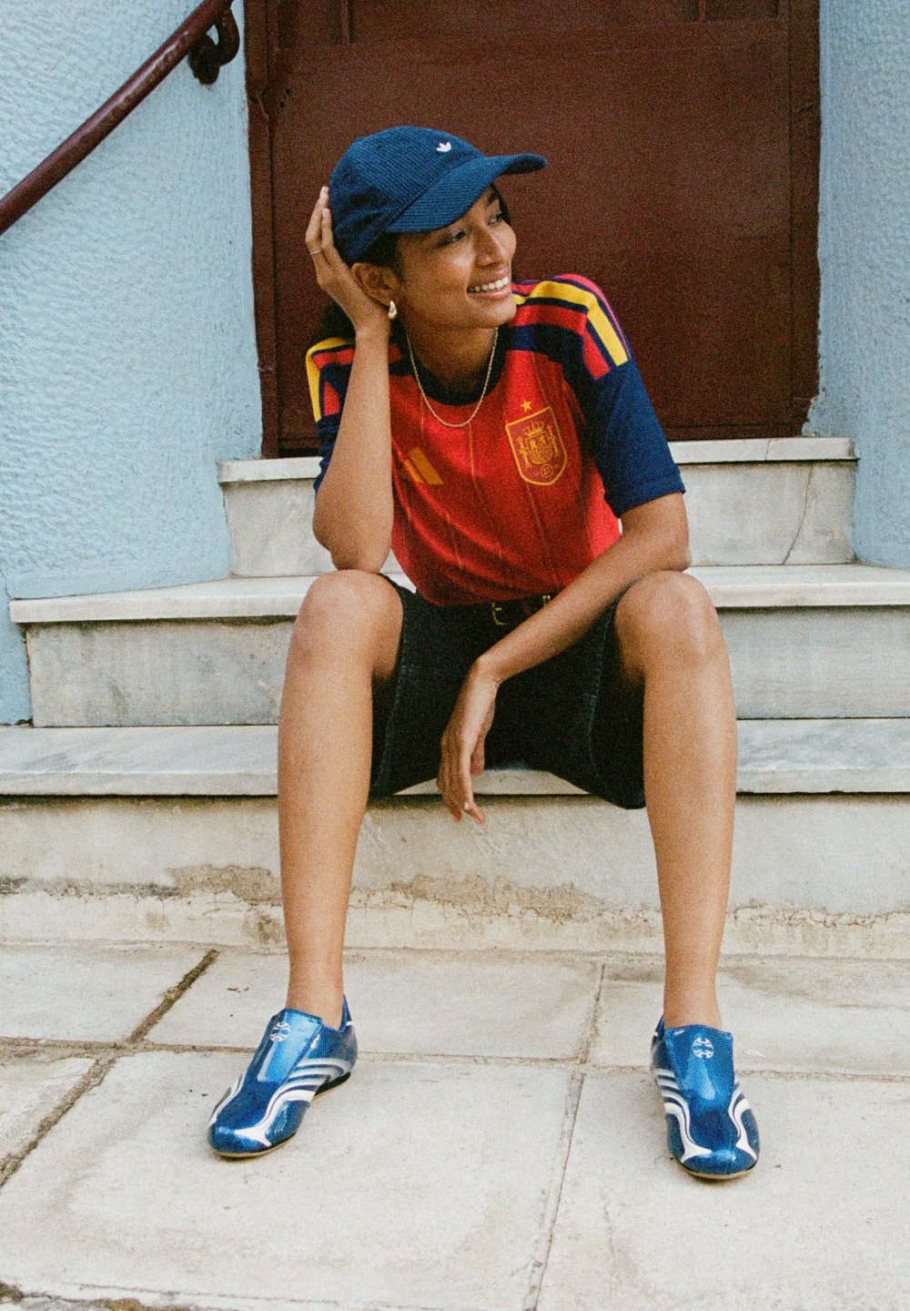 Νέος άτομο κάθεται σε τσιμεντένια σκαλιά, φορώντας αθλητική φανέλα σε κόκκινο και μπλε χρώμα, μπλε καπέλο, μαύρη βερμούδα και μπλε αθλητικά παπούτσια, χαμογελώντας και κοιτάζοντας δεξιά.