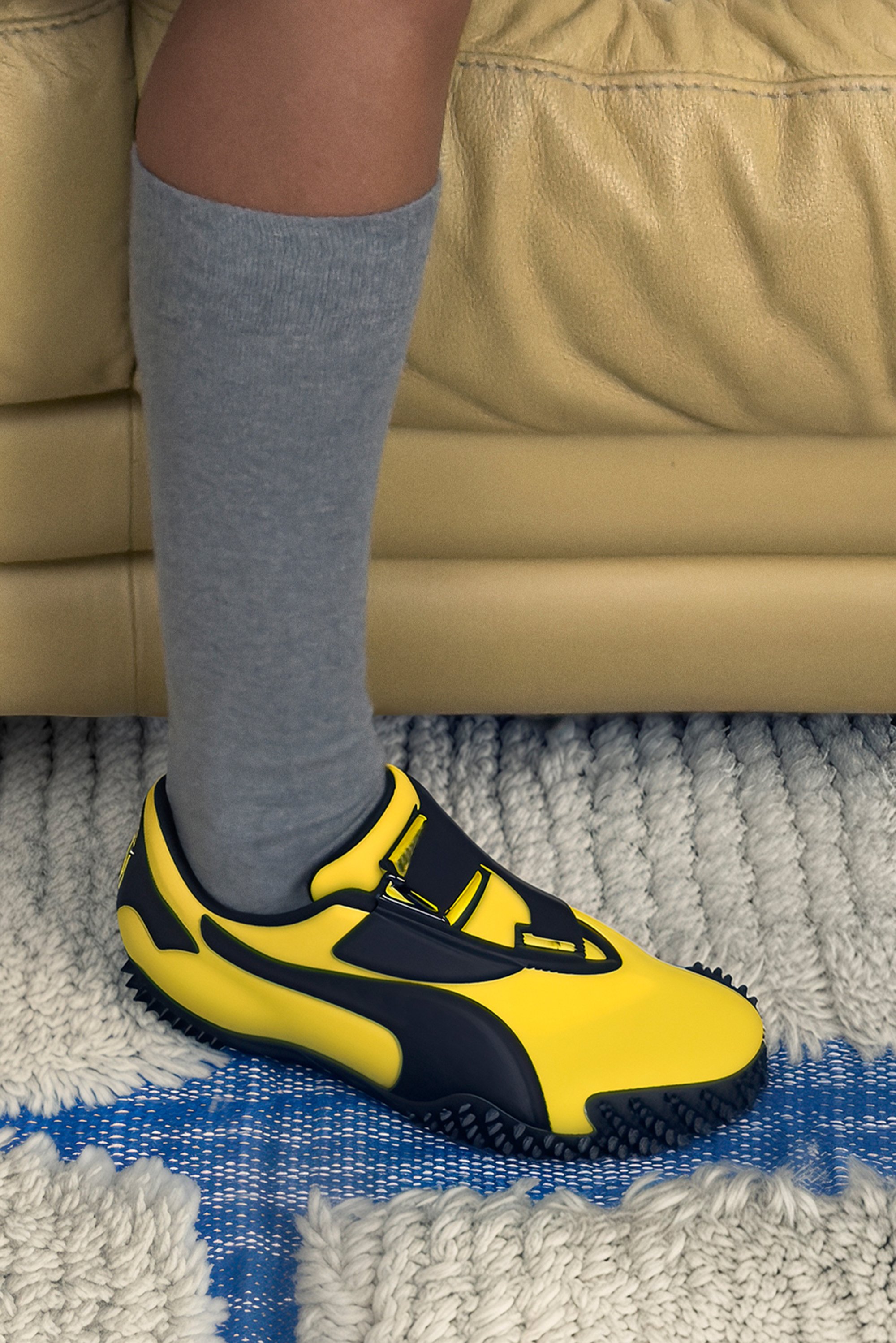 Zapatilla deportiva amarilla y negra con suela de goma y dos correas, combinada con una media gris hasta la rodilla, sobre una alfombra blanca texturizada.