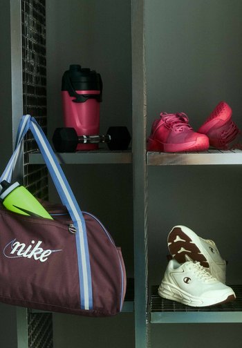Sac de sport Nike bordeaux avec bouteille d'eau, haltère, shaker rose, chaussures de sport rouges et blanches sur des étagères métalliques dans un vestiaire.