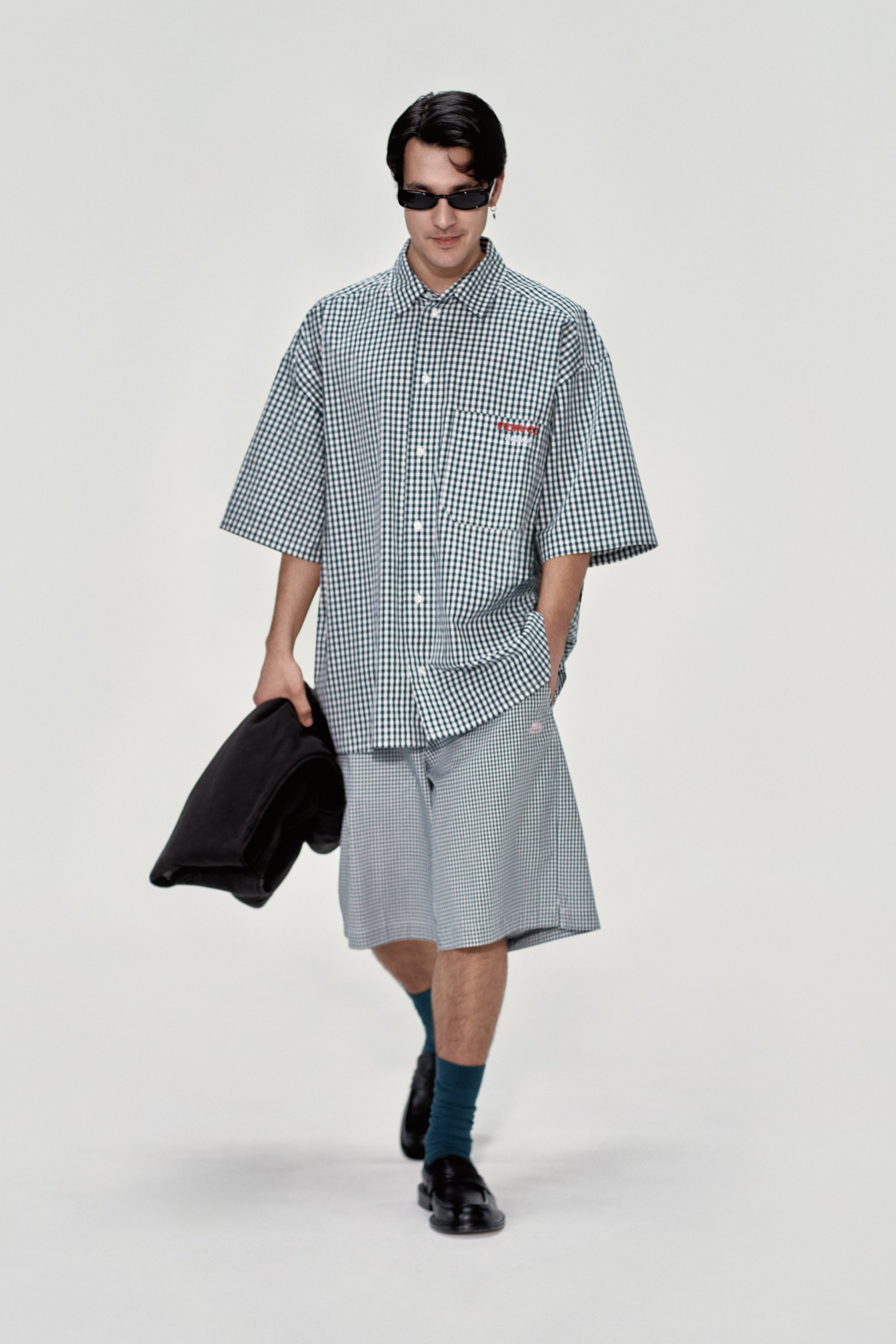 Giovane uomo cammina con sicurezza, indossando una camicia a quadri oversized e pantaloni corti, con in mano una giacca nera, occhiali da sole e calze turchesi.