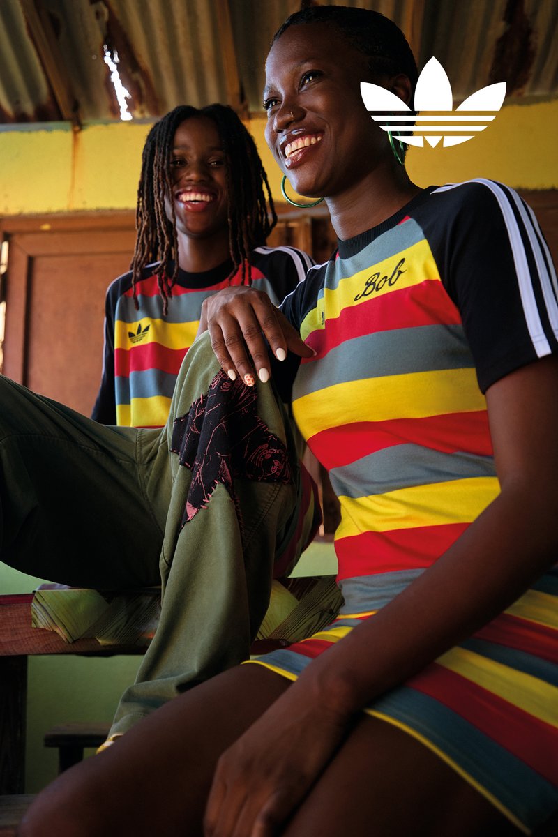 Two smiling individuals wearing striped Adidas clothing in an indoor rustic setting, one resting a hand on their knee while holding a jacket.
