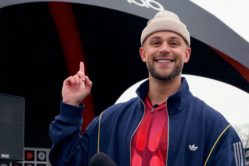 Smiling man wearing a beige beanie and blue jacket, pointing upward with his right hand at a black and red structure behind him.