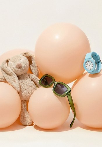 Plišasta bež zajček, zelena sončna očala in modra ročna ura, ki počivajo na velikih breskovo obarvanih balonih na svetlem ozadju.