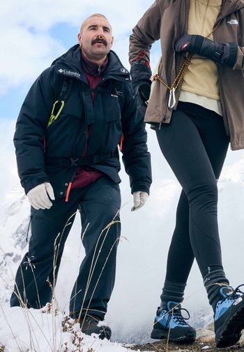 Δύο αναρριχητές με χειμερινά ρούχα και ιμάντες αναρρίχησης περπατούν σε χιονισμένο βουνό κάτω από έναν μερικώς συννεφιασμένο ουρανό.