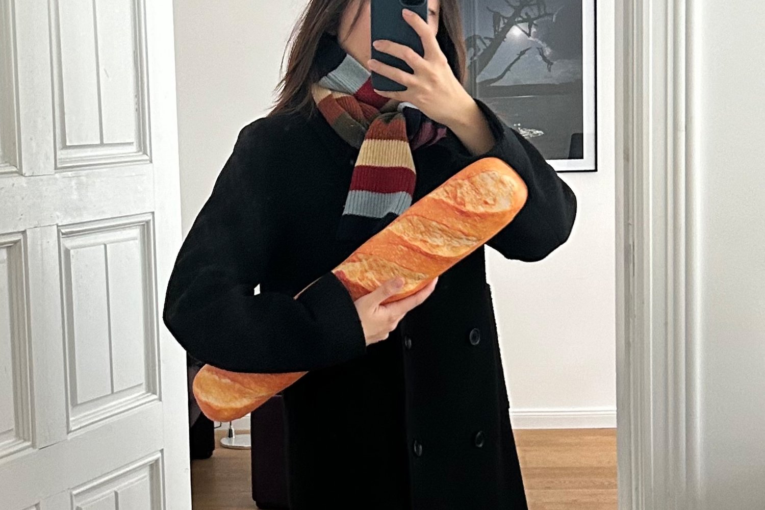 Een vrouw in een zwarte jas houdt een baguette vast en maakt een selfie, gekleed in een kleurrijke gestreepte sjaal, met een effen achtergrond.