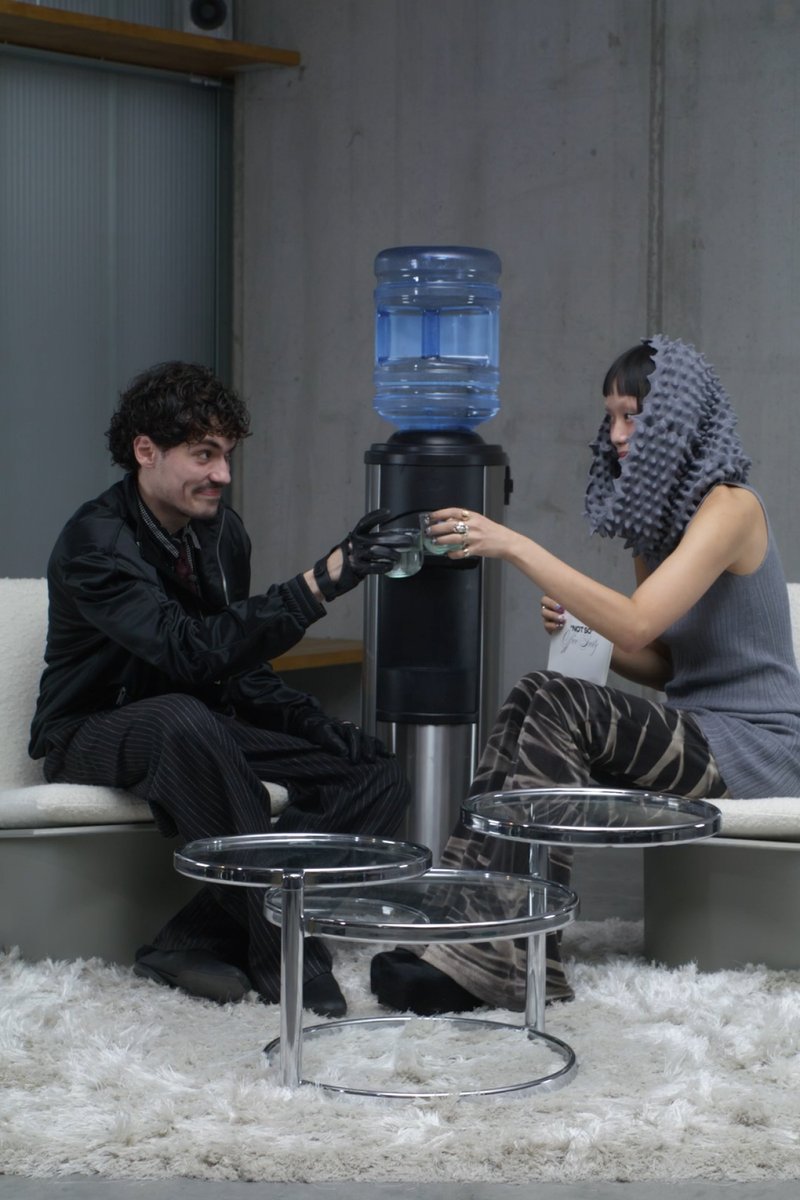 Ein Mann in schwarzen Handschuhen und gestreiften Hosen teilt sich ein Glas mit einer Frau in einem grauen Outfit und einer auffälligen Kopfbedeckung an einem Wasserspender.