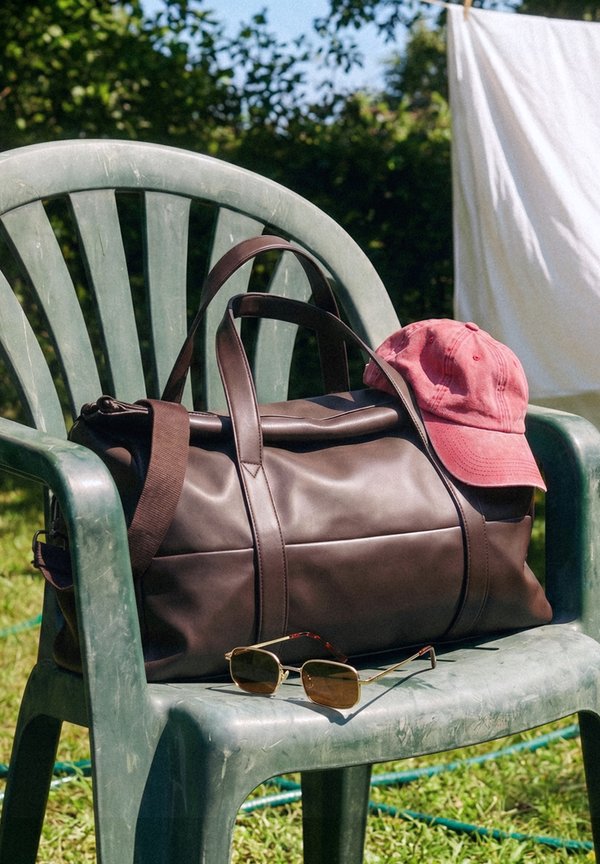 Bolsa de lona de cuero con una gorra de béisbol roja encima y gafas de sol en una silla de plástico verde al aire libre con césped y ropa tendida al fondo.