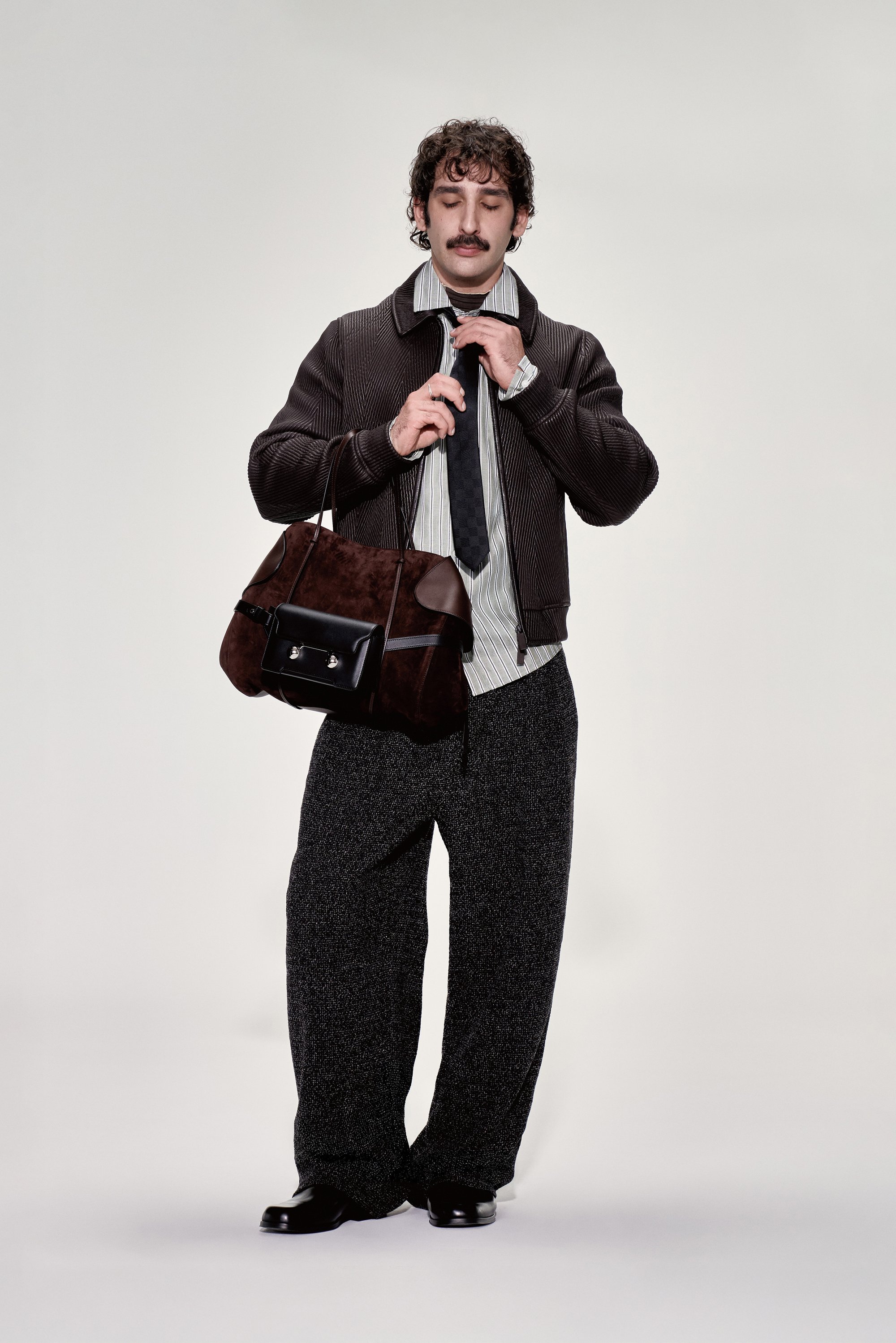 Un hombre con un atuendo elegante ajusta su corbata mientras sostiene una bolsa marrón. Su vestimenta incluye una chaqueta oscura y pantalones grises holgados.